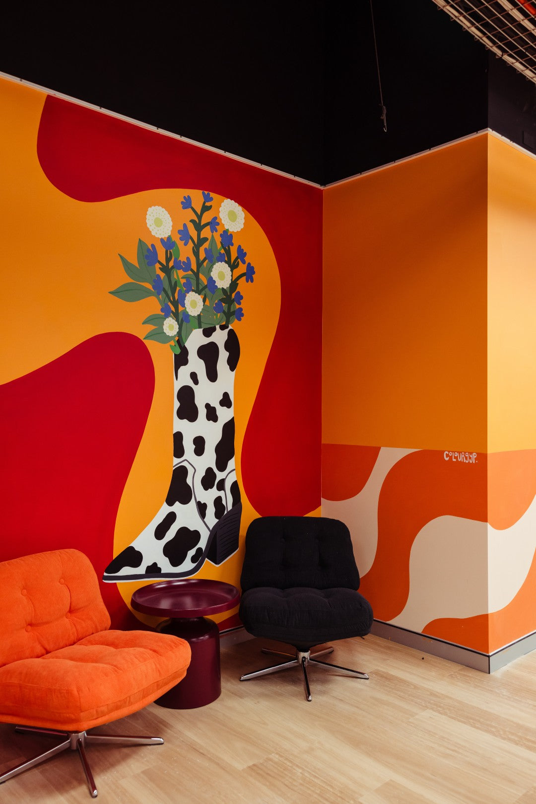 A modern interior corner with bold abstract wall art in orange, red, and cream, featuring a stylized black-and-white boot with flowers. The room includes an orange upholstered chair, a black swivel chair, a small dark side table, and light wood flooring.